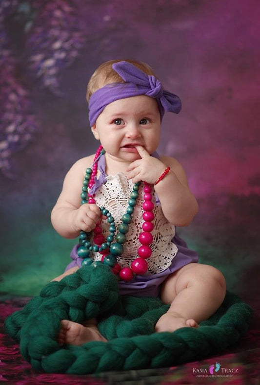 A baby girl sitting on a dark green chunky knit blanket, wearing a purple lace top, a purple headband, and colorful green and pink beaded necklaces against a purple and green studio backdrop.