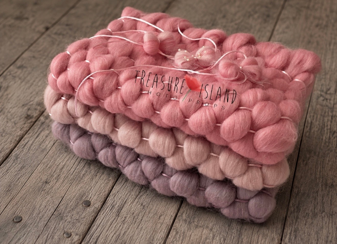 A stack of three  Fluffy Marshmallow Blankets in dusty pink, pale blush, and mauve, wrapped in clear cellophane with a small pink bead and the text 'Treasure Island Hand Knit' visible on the top blanket, set on a rustic wooden surface.