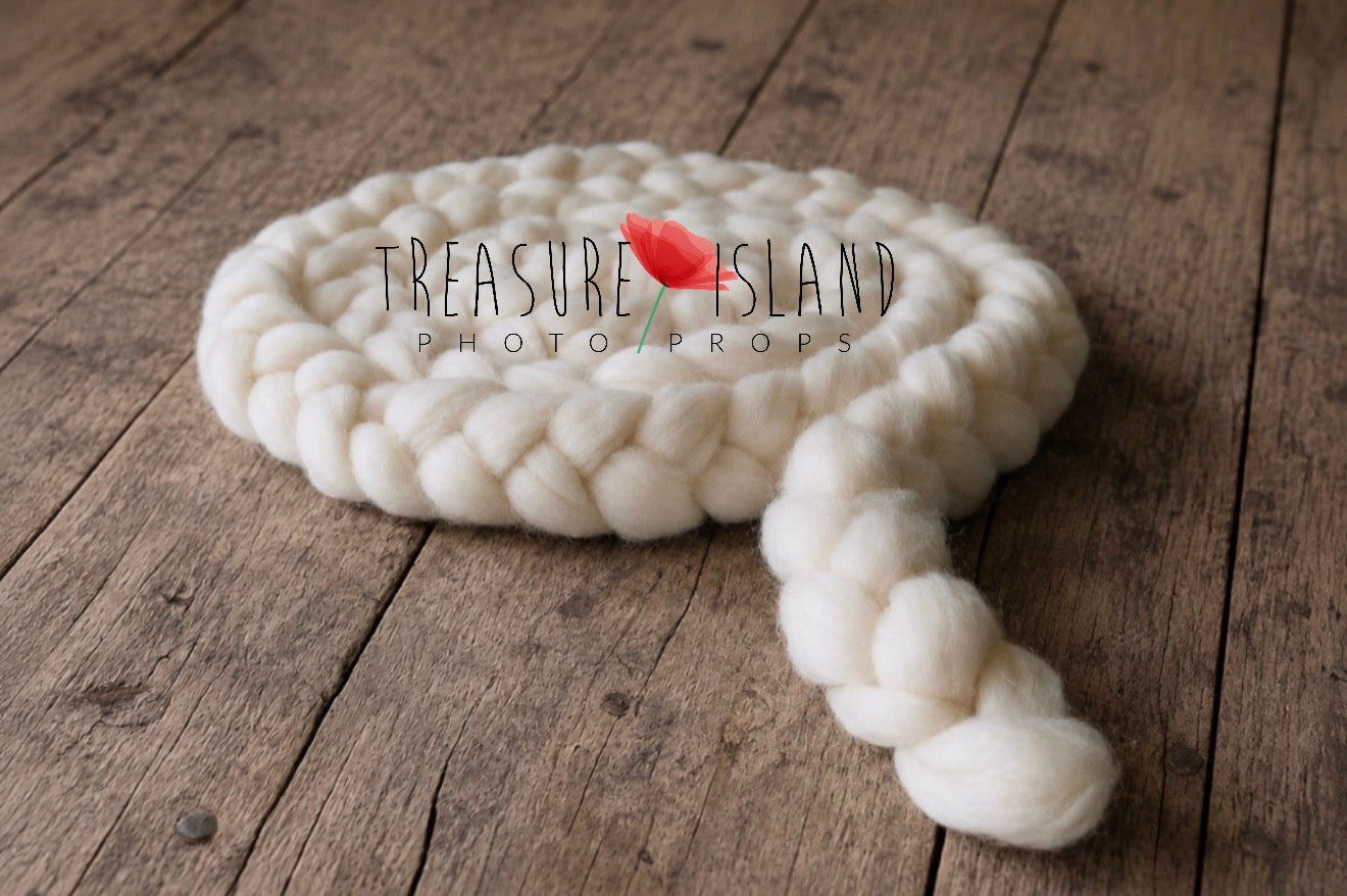 A chunky braided knit blanket in off-white is coiled on a rustic wooden surface. A small red flower prop is placed on top of the blanket, and a watermark reading 'TREASURE ISLAND PHOTO PROPS' is overlaid across the center.