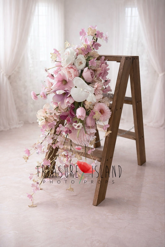 Floral photography prop arrangement with pink and blush flowers cascading over rustic wooden ladder, soft romantic studio background
