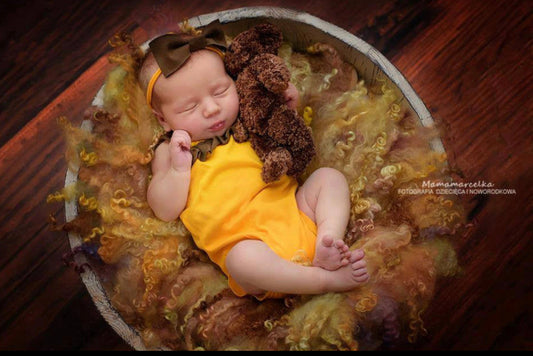 Newborn baby in a yellow outfit with a brown bow and teddy bear, lying on a textured surface.