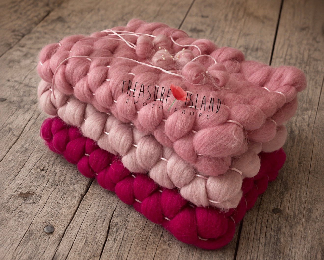 A stack of chunky knitted wool blankets in dusty pink, light pink, and bright magenta, placed on a rustic wooden surface. A small red heart logo with the text 'Treasure Island Photo Props' is visible on the top blanket.