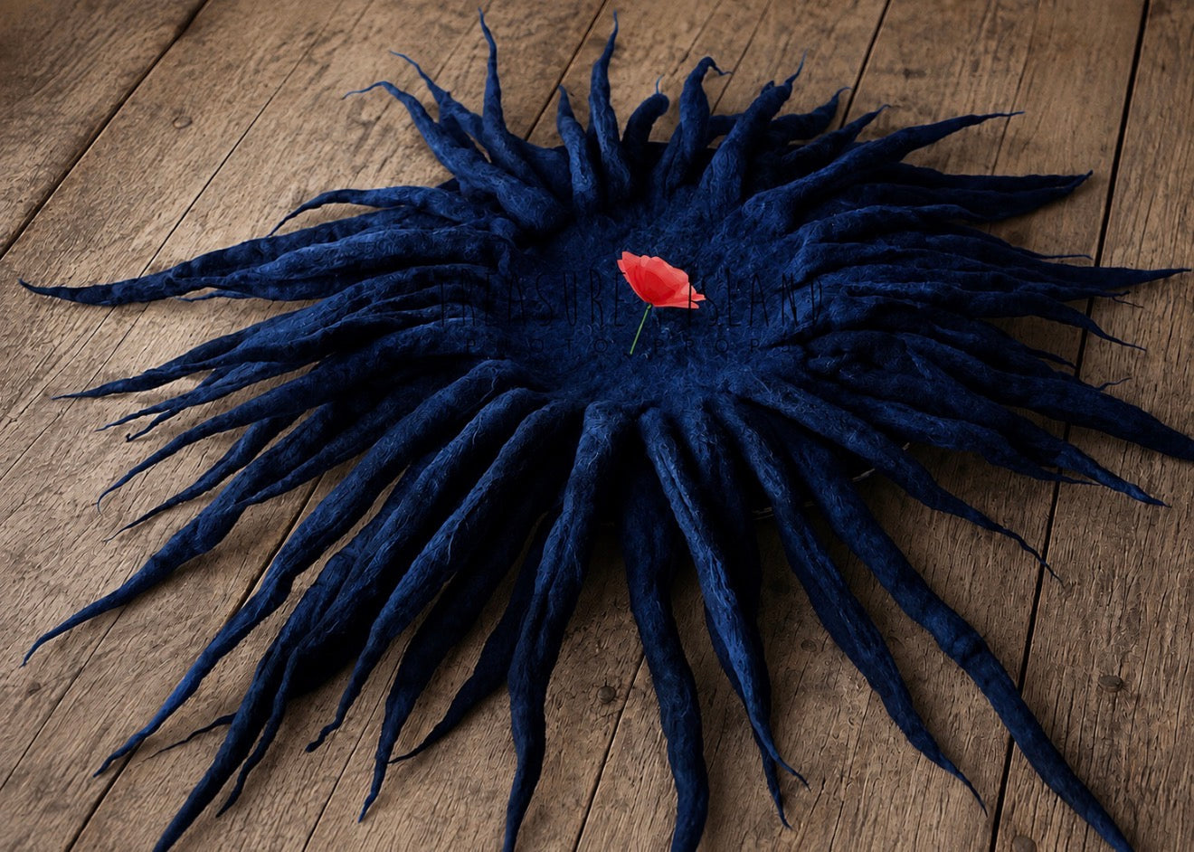 A dark navy blue felted wool sunburst or dreads-style textile piece laid flat on a rustic wooden surface, with a small red flower placed in the center.