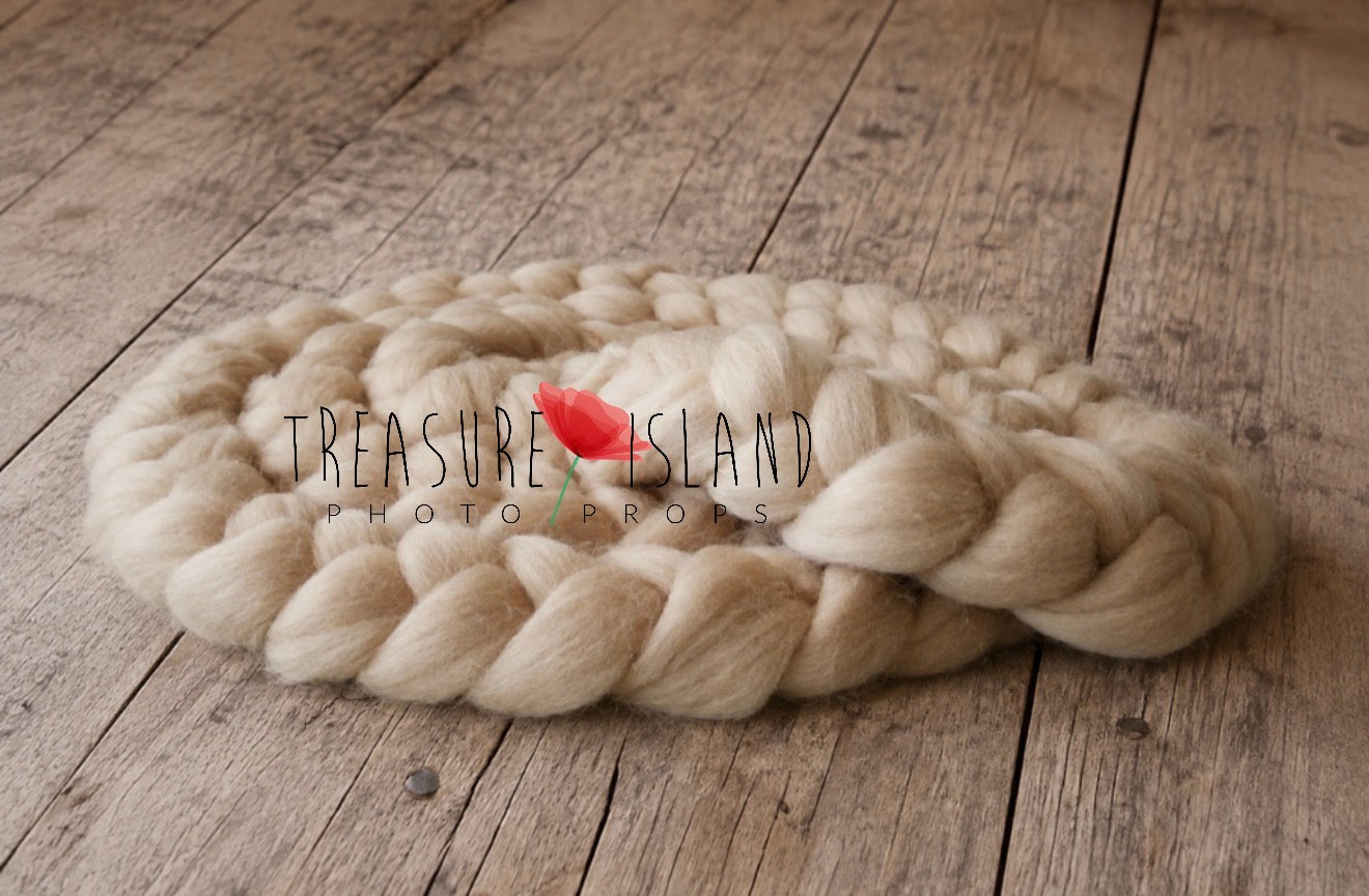 A large, chunky braided wool rug in a light oatmeal beige color, styled on a rustic wooden plank floor with a small red tulip flower accent in the center. A watermark for 'Treasure Island Photo Props' is visible across the middle.