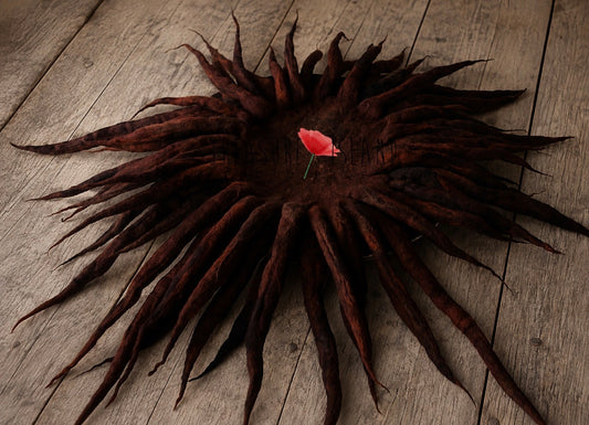 A dark brown dreadlock hairpiece arranged in a circular, sunburst shape on a rustic wooden surface, with a small red flower tucked into the center.