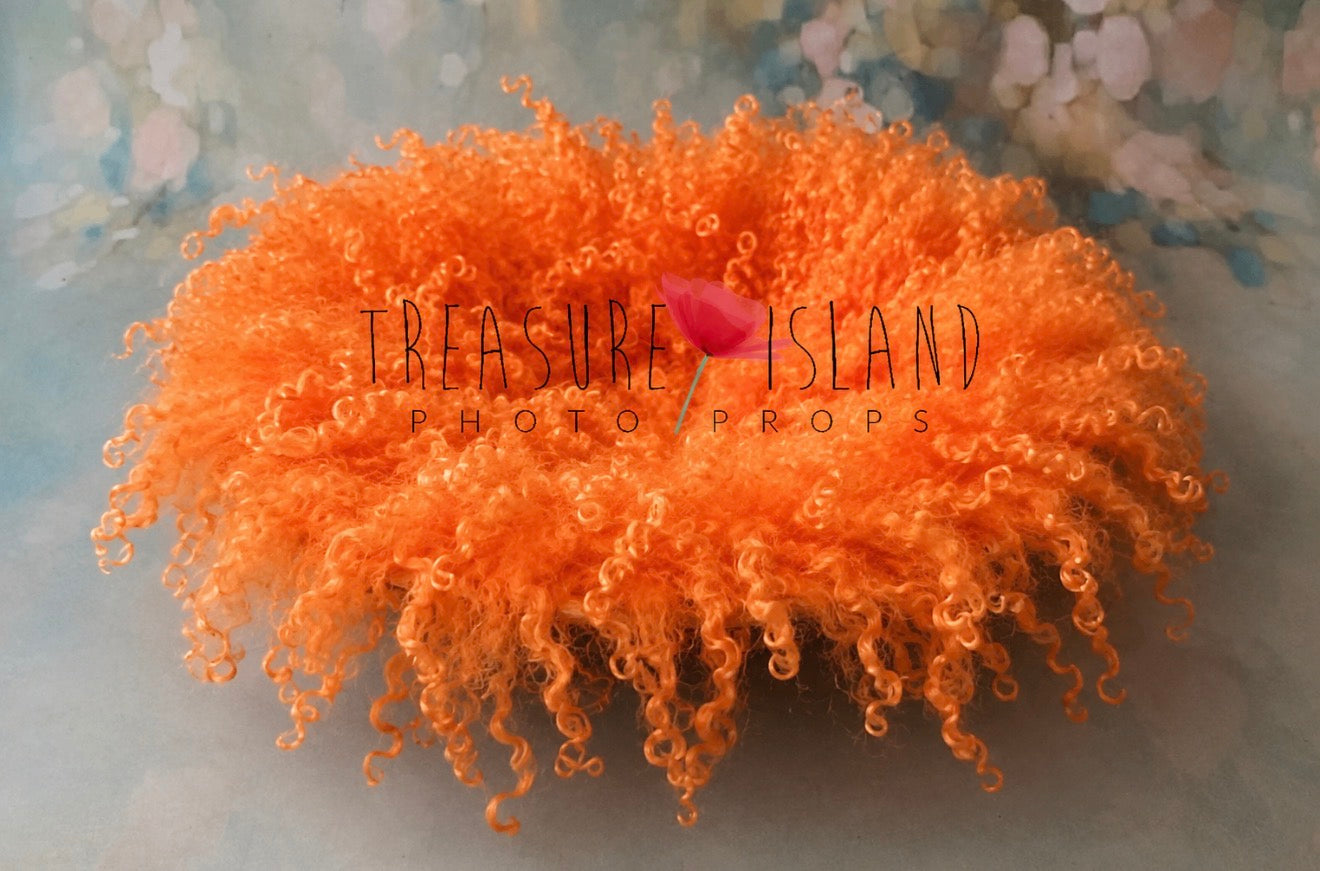 A bright orange curly wig displayed on a soft, blurred pastel background with a small pink flower prop in the center. The text 'Treasure Island Photo Props' is overlaid across the middle.