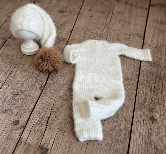 Cream fluffy knitted newborn photography romper with long sleeves and legs, displayed beside a matching elongated hat with a hazelnut-brown pom-pom on rustic wooden flooring.