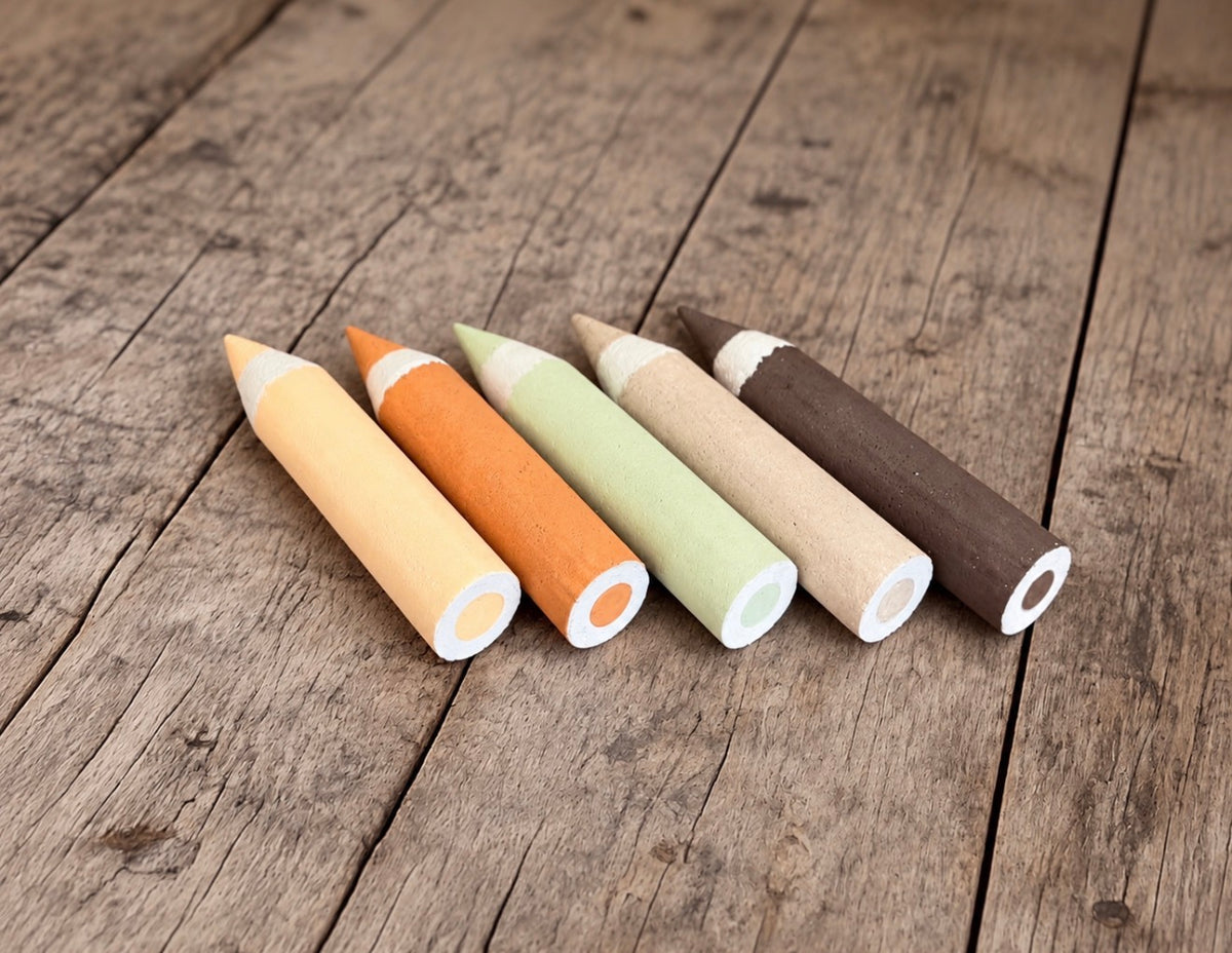 Five oversized styrofoam crayon photography props in yellow, orange, mint green, beige and dark brown, arranged on a rustic wooden floor.