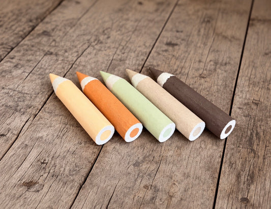 Five oversized styrofoam crayon photography props in yellow, orange, mint green, beige and dark brown, arranged on a rustic wooden floor.