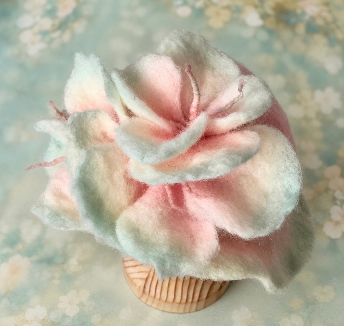 Hand-felted newborn flower hat shown from the front, featuring layered pink and pale green wool petals and hand-dyed edges