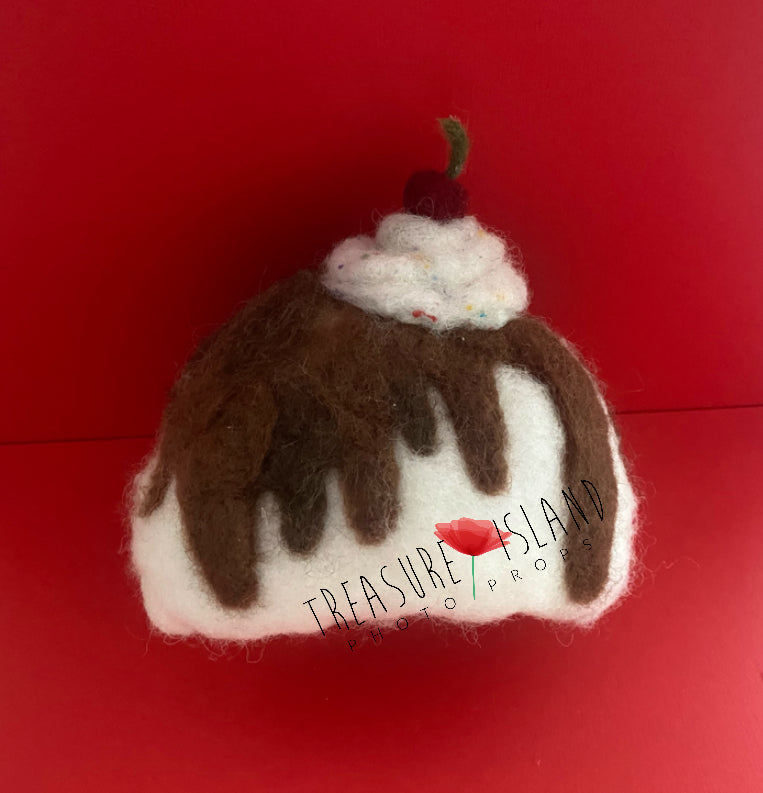 A handmade hat designed to look like a cupcake, with a brown and white color scheme, topped with a fake cherry, displayed against a red background.
