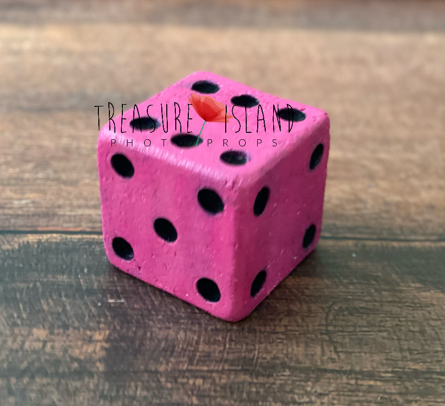 Single handmade pink styrofoam dice with black dots and a textured finish, displayed on rustic wooden flooring