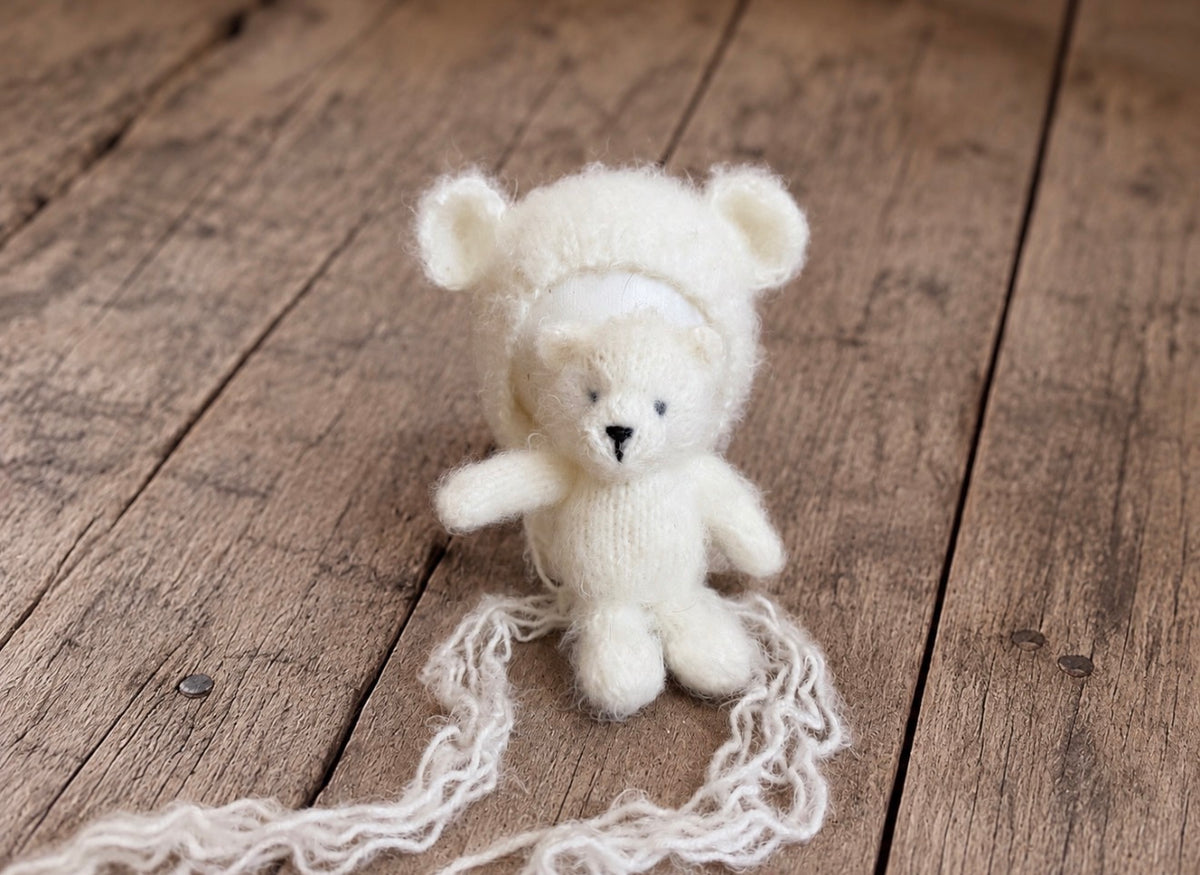 Miniature knitted teddy bear displayed with a matching fluffy bear-ear newborn bonnet and long decorative ties on a wooden background.
