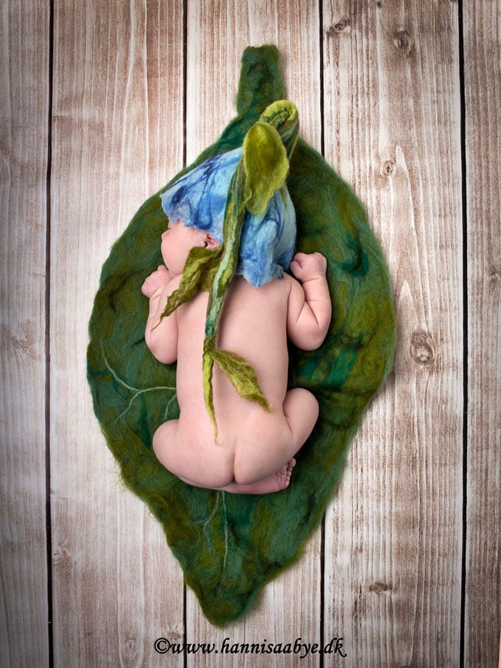 Newborn wearing a blue felted flower hat with long stalk posed on a large hand-felted green leaf